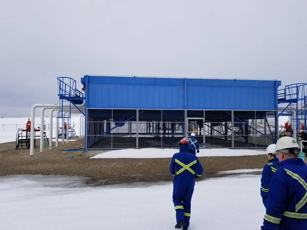 Crew in coveralls and hard hats walking out to a blue air cooler at a snowy gas site